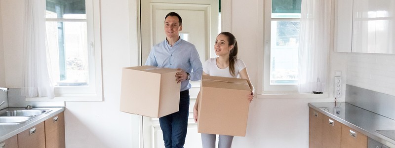 a couple holding cardboard boxes walking inside a kitchen