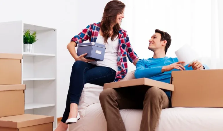 A couple is sitting on sofa with moving boxes all around.